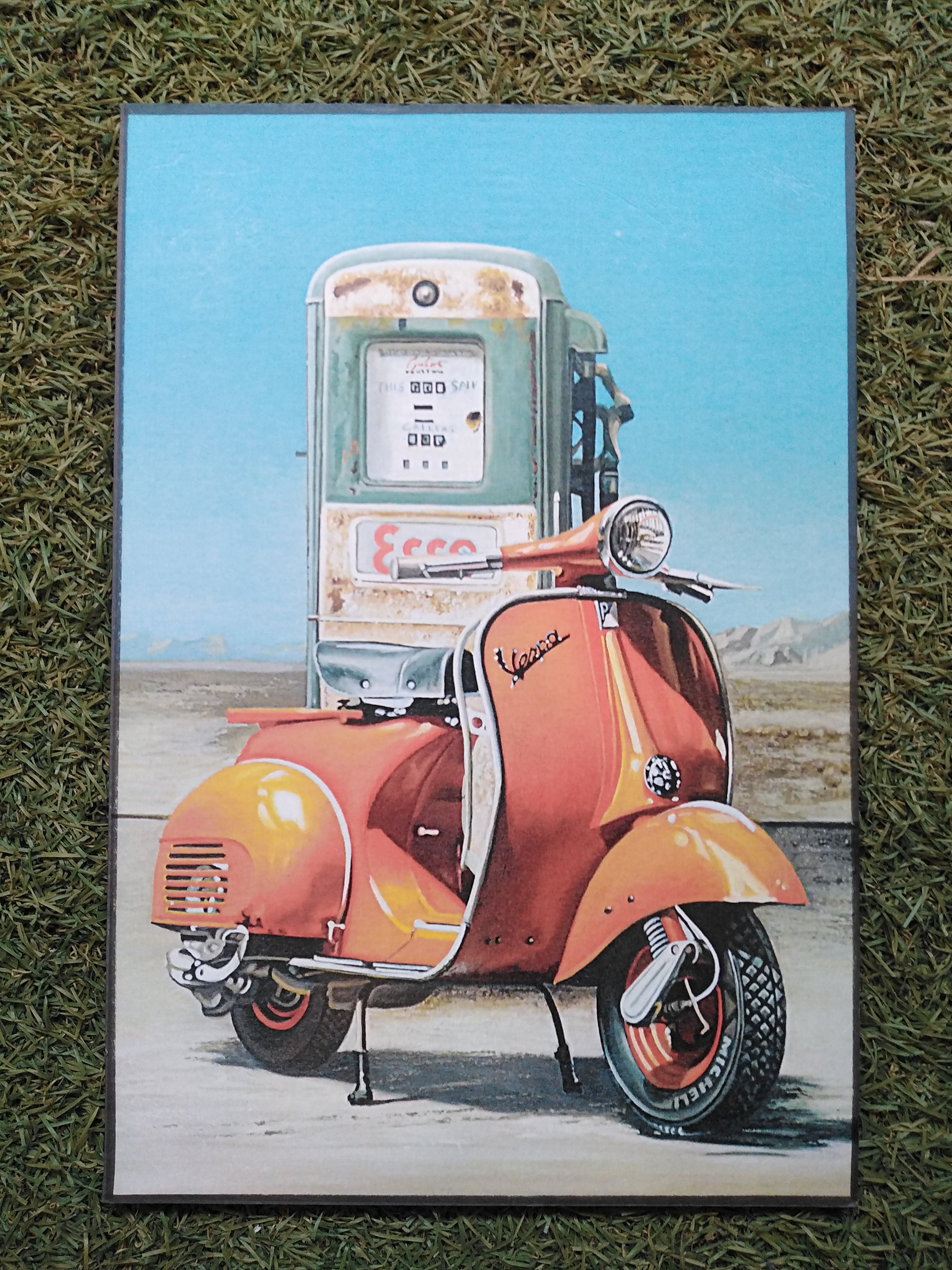 Cuadro de madera Vespa naranja clásica aparcada frente a gasolinera Esso oxidada bajo cielo azul con montañas