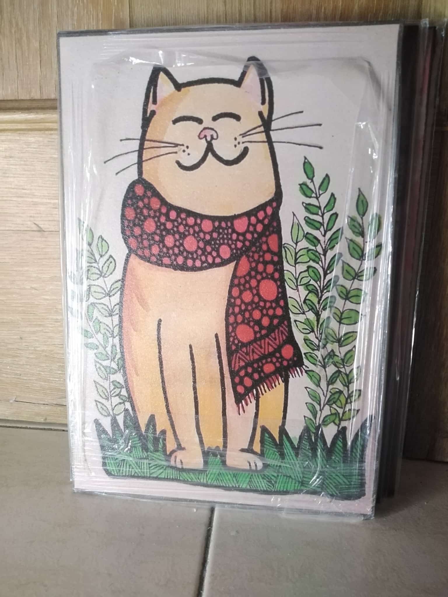 Cuadro ilustración gato crema sonriente con bufanda lunares rojos entre plantas verdes madera 19x28 cm